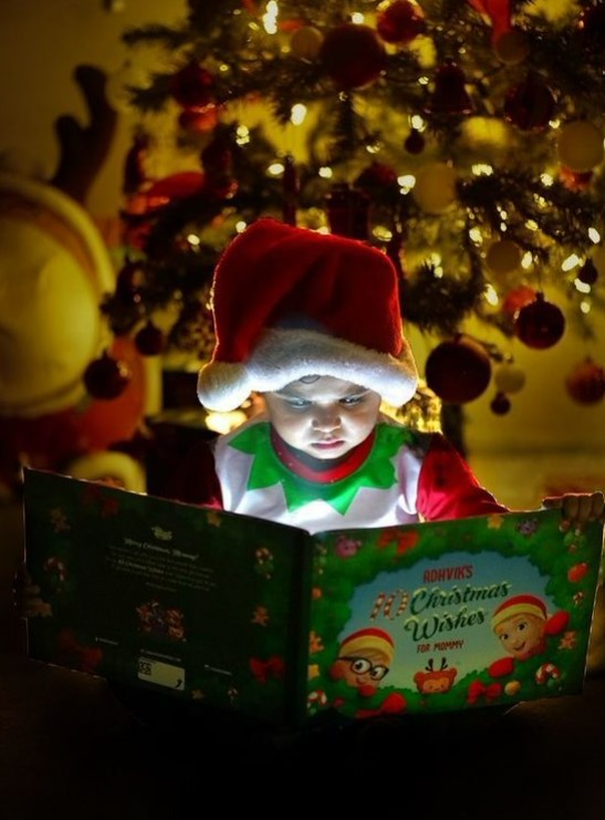 A child dressed as an elf reads his own personalized Christmas book for Mommy and child in front of an xmas tree.