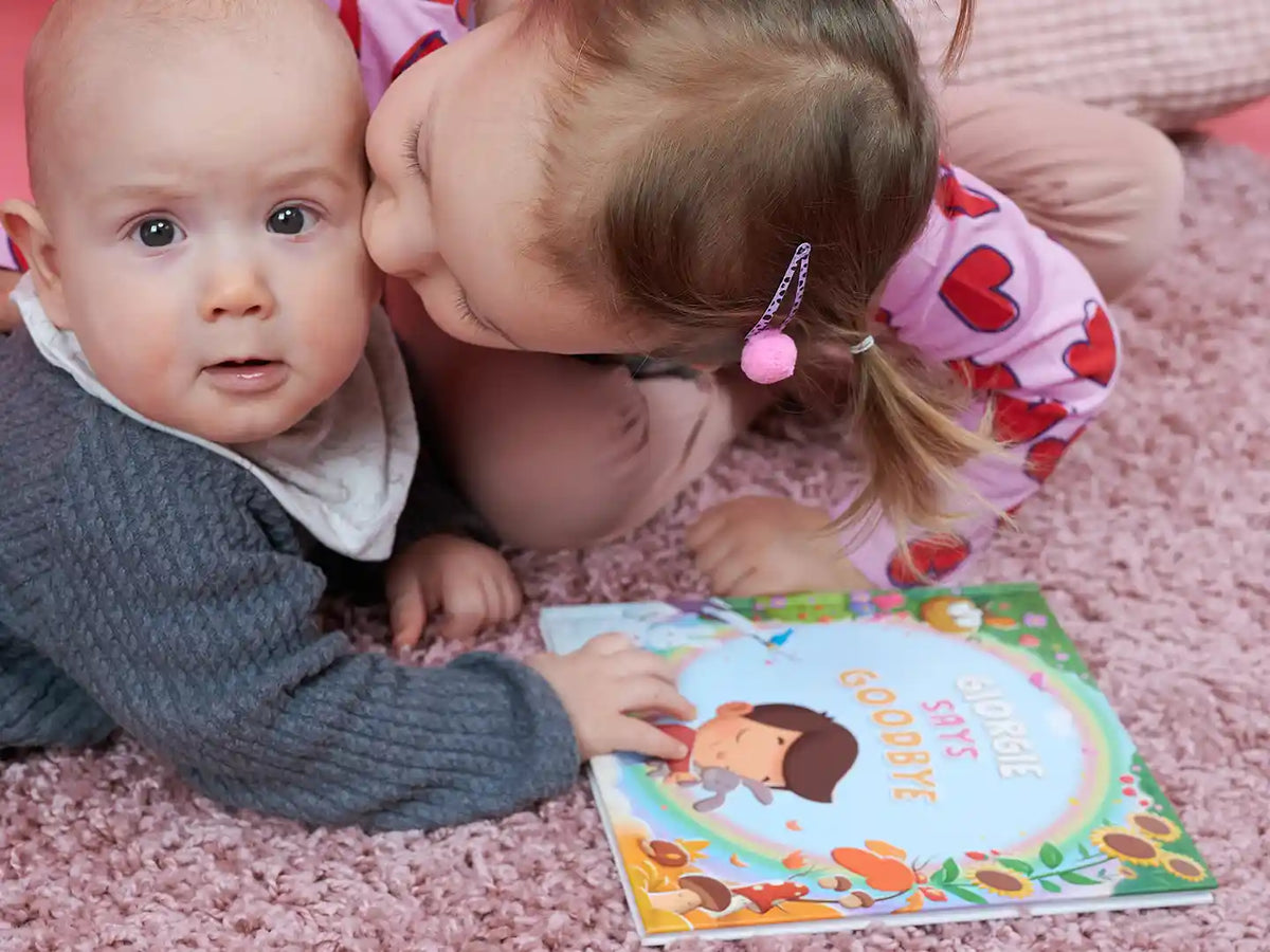 Girl kissing her little brother, with a personalized book about healing after loss lying beside them.