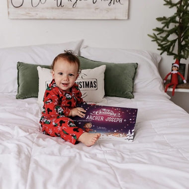 A little boy smiles on a bed holding his custom Christmas book for one kiddo.